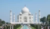 The Taj Mahal at sunrise, with its iconic white marble reflecting in the tranquil waters of the garden's pool. Visitors admire the stunning architecture surrounded by lush green gardens.