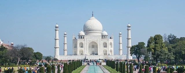 The Taj Mahal at sunrise, with its iconic white marble reflecting in the tranquil waters of the garden's pool. Visitors admire the stunning architecture surrounded by lush green gardens.