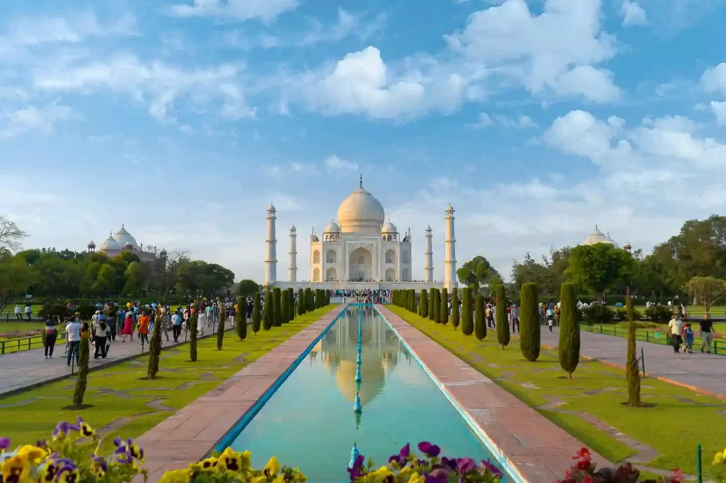 The Taj Mahal at sunrise, with its iconic white marble reflecting in the tranquil waters of the garden's pool. Visitors admire the stunning architecture surrounded by lush green gardens.