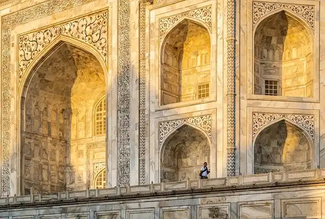 The Taj Mahal at sunrise, with its iconic white marble reflecting in the tranquil waters of the garden's pool. Visitors admire the stunning architecture surrounded by lush green gardens.