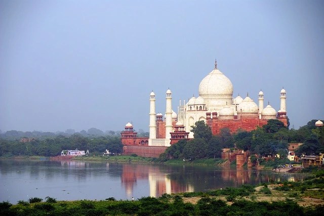 The Taj Mahal at sunrise, with its iconic white marble reflecting in the tranquil waters of the garden's pool. Visitors admire the stunning architecture surrounded by lush green gardens.