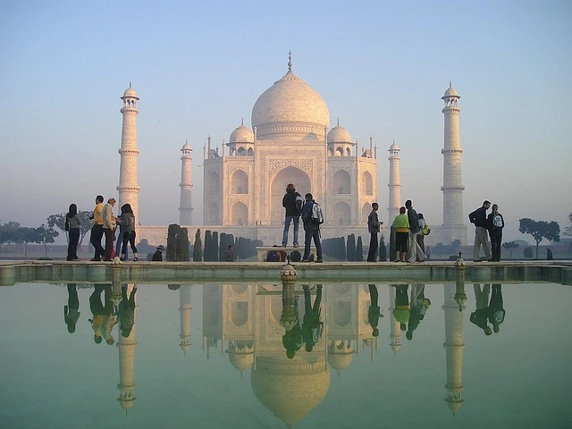 The Taj Mahal at sunrise, with its iconic white marble reflecting in the tranquil waters of the garden's pool. Visitors admire the stunning architecture surrounded by lush green gardens.