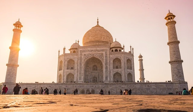 The Taj Mahal at sunrise, with its iconic white marble reflecting in the tranquil waters of the garden's pool. Visitors admire the stunning architecture surrounded by lush green gardens.