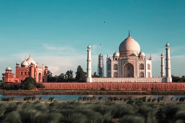 The Taj Mahal at sunrise, with its iconic white marble reflecting in the tranquil waters of the garden's pool. Visitors admire the stunning architecture surrounded by lush green gardens.