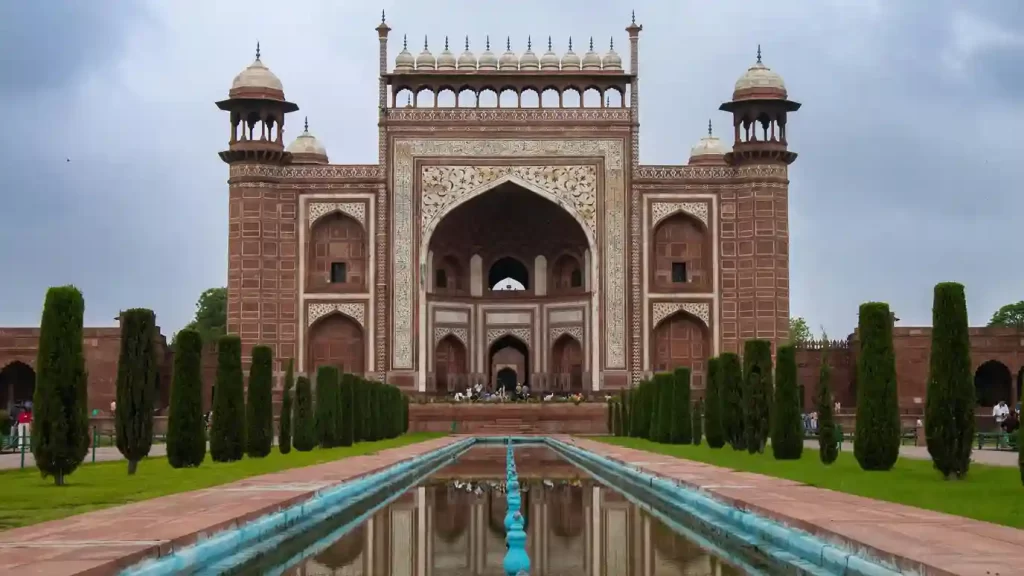 The Taj Mahal at sunrise, with its iconic white marble reflecting in the tranquil waters of the garden's pool. Visitors admire the stunning architecture surrounded by lush green gardens.