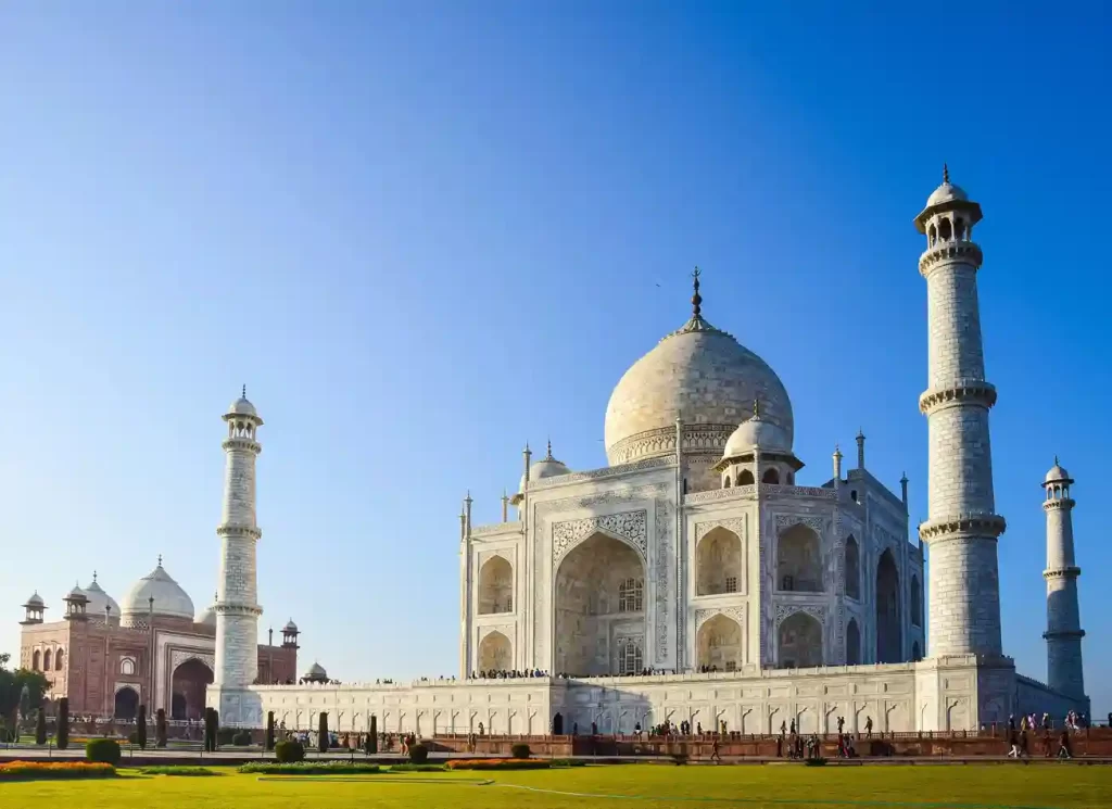 The Taj Mahal at sunrise, with its iconic white marble reflecting in the tranquil waters of the garden's pool. Visitors admire the stunning architecture surrounded by lush green gardens.