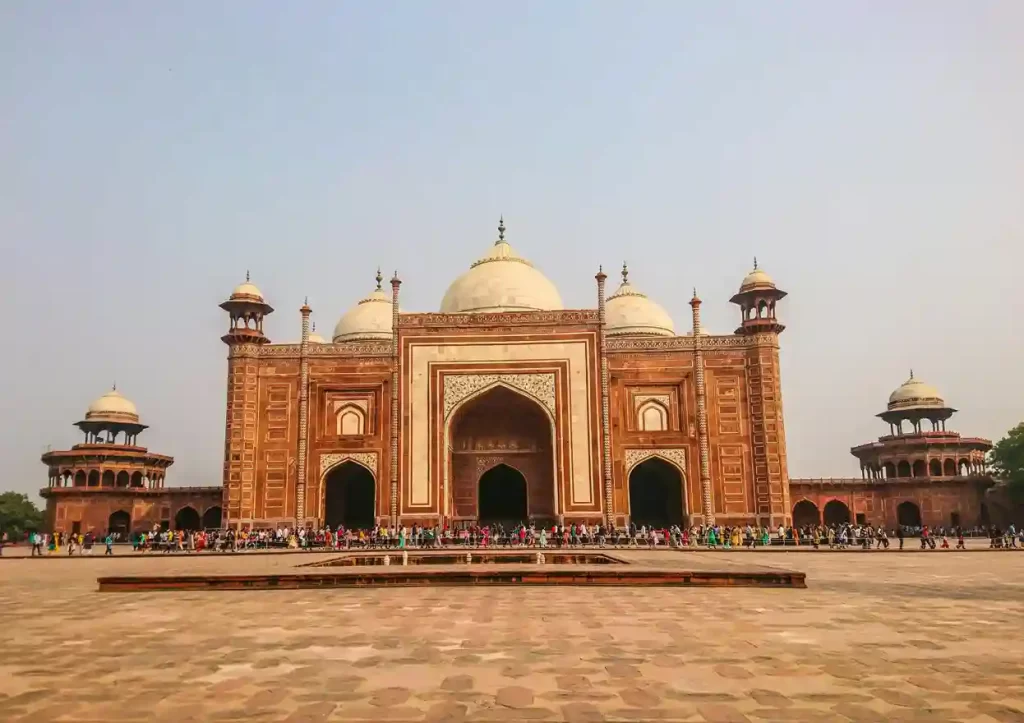 The Taj Mahal at sunrise, with its iconic white marble reflecting in the tranquil waters of the garden's pool. Visitors admire the stunning architecture surrounded by lush green gardens.