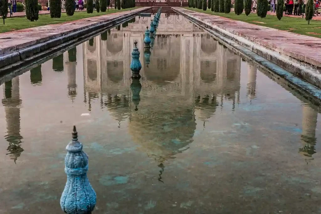 The Taj Mahal at sunrise, with its iconic white marble reflecting in the tranquil waters of the garden's pool. Visitors admire the stunning architecture surrounded by lush green gardens.